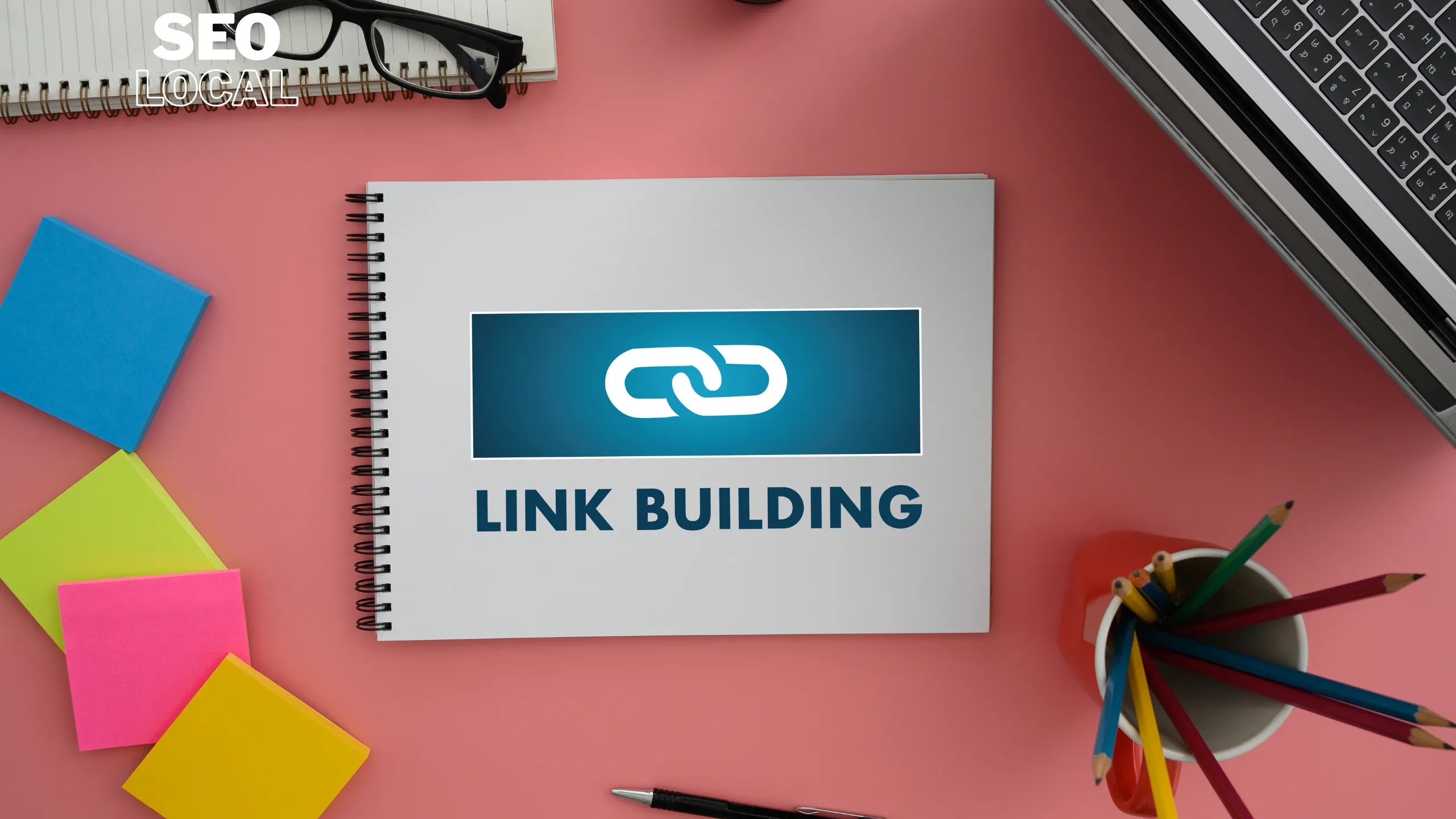Un cuaderno sobre una mesa rosa con el icono de un eslabón y las palabras link building, junto a notas de colores y lápices